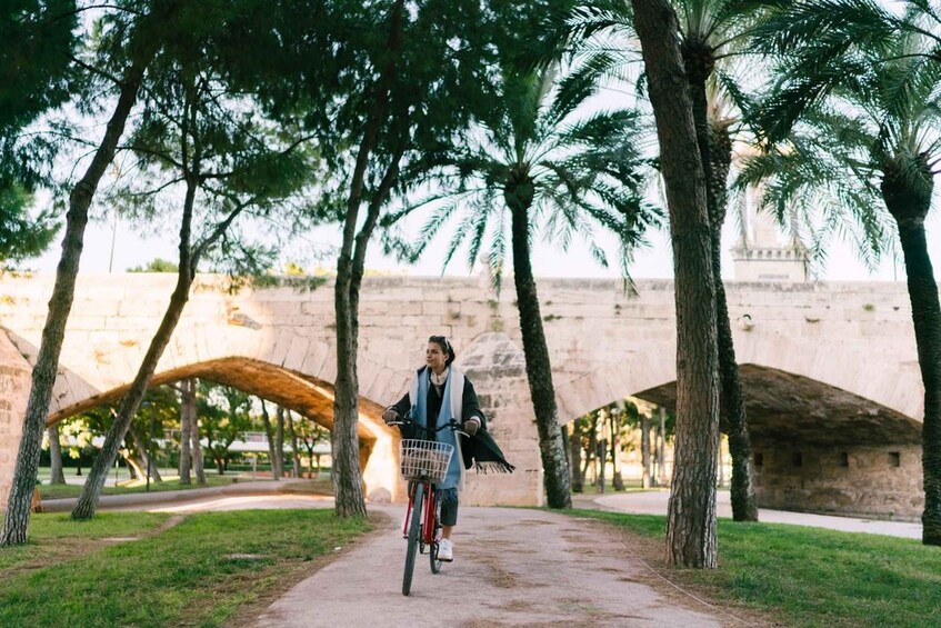 A person riding a bicycle on a paved path surrounded by trees and a stone bridge. 2