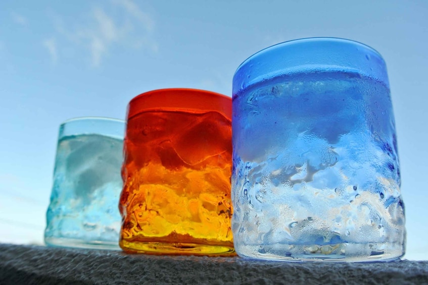 Three glass containers with different colored liquids, placed on a textured surface against a clear sky. 1
