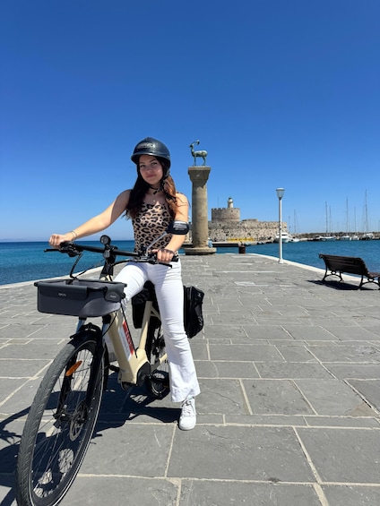 A person on a stationary electric bike with a helmet, white pants, and a leopard print top, standing on a paved area with a stone wall and a statue in the background. 2