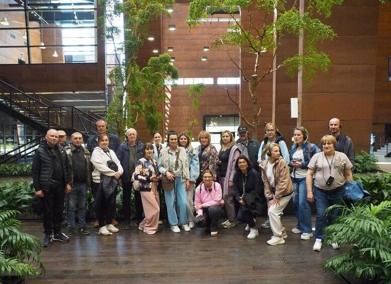 A group of people standing in an indoor area with wooden flooring, surrounded by green plants and a modern building with large windows in the background. 2