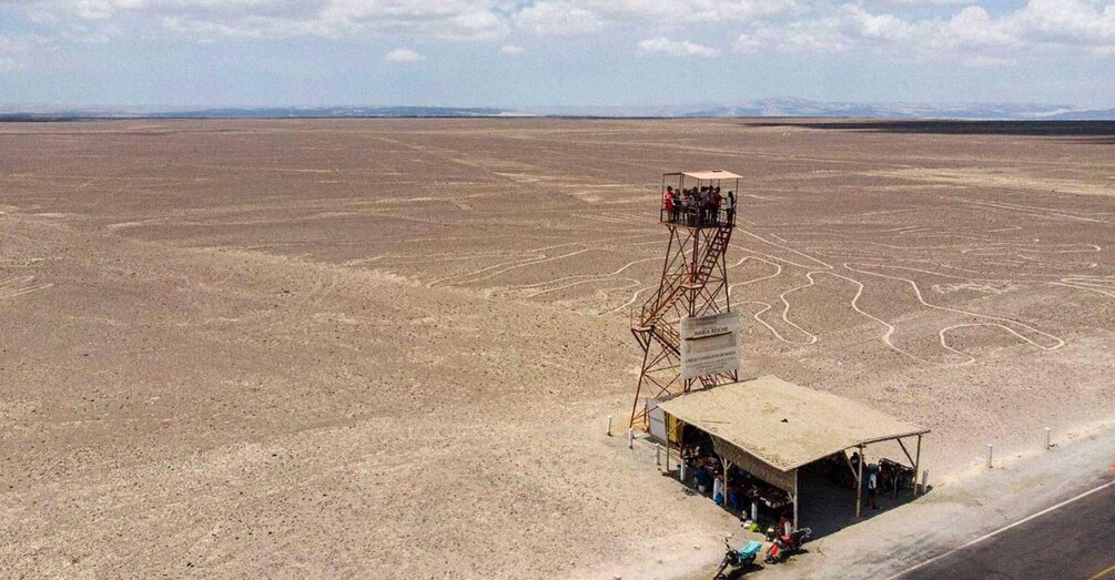 A desert landscape with a tall metal tower and a small building with people around it. 4