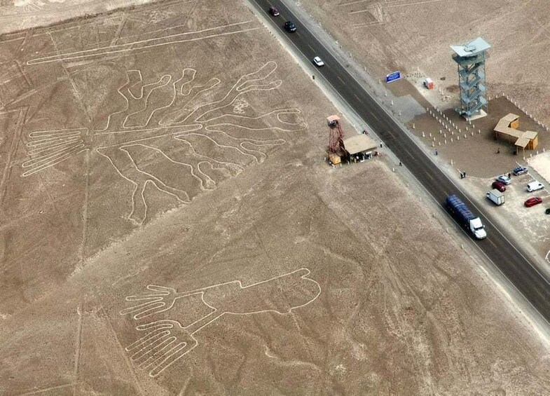 An aerial view of a desert landscape with large handprint-like rock formations. A road runs parallel to the formations, with vehicles and a small structure nearby. 5