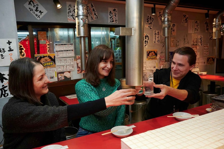 Three people toasting with drinks at a restaurant with Japanese menu boards. 1