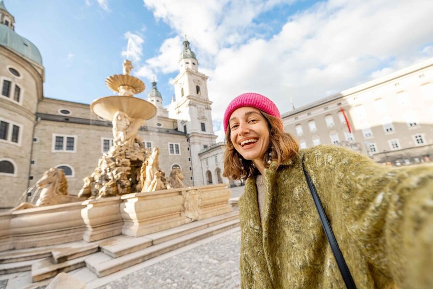 A person in a green coat and pink beanie standing in front of a historic fountain with statues and a building with domes in the background. 2