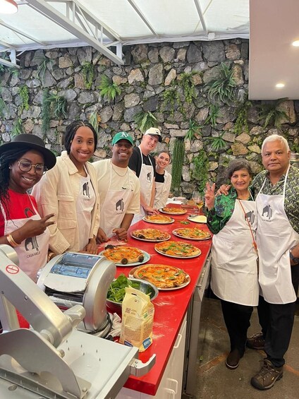 A group of people in a kitchen with stone walls and plants, preparing pizzas. 1