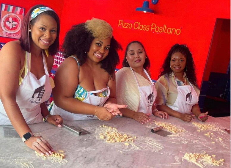 Four people at a pizza-making station with a red background and a sign that reads 'Pizza Class Positano.' 4