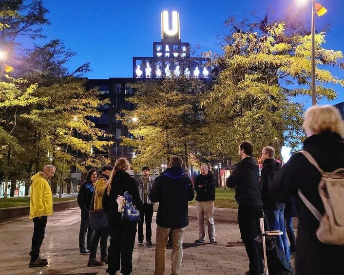 A group of people standing on a paved path at night, with a building featuring a prominent illuminated 'U' in the background. 1