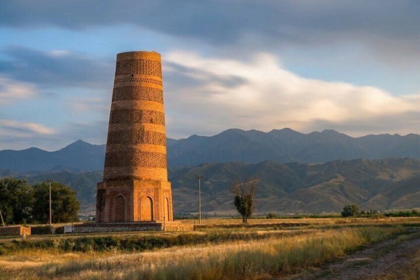 A tall, cylindrical tower with a square base, surrounded by a grassy field and mountains in the background. 1