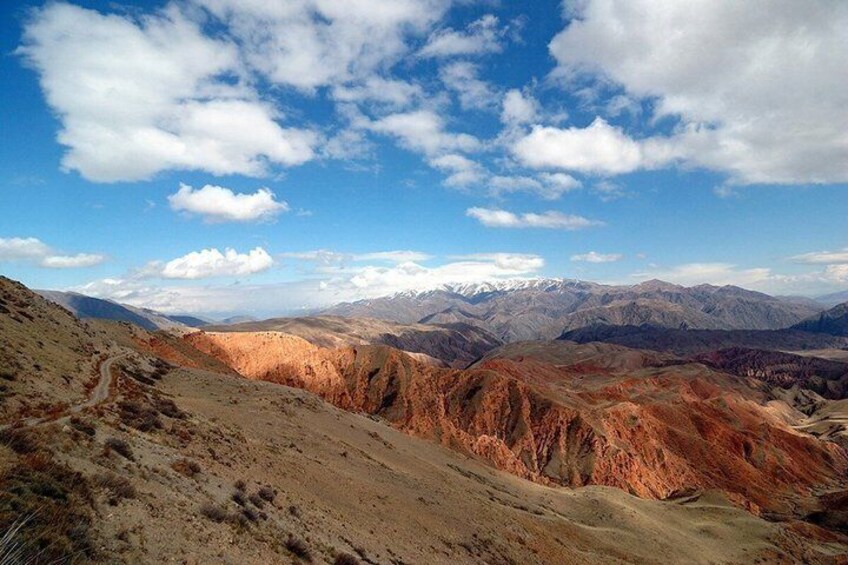 A mountainous landscape with a dirt path, distant snow-capped peaks, and a clear blue sky. 2