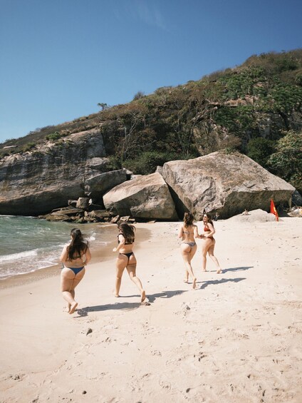 Three women walking on a sandy beach with large rocks and cliffs in the background. 1