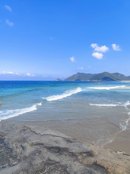 A beach with waves, a rocky foreground, and distant mountains under a clear sky. 4