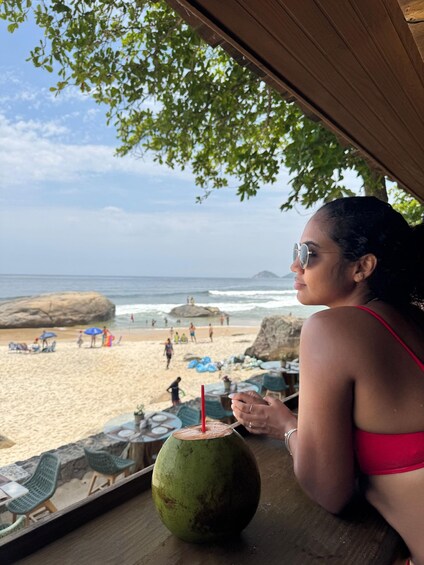 A person in a red swimsuit sitting at a wooden table with a green coconut drink. The background features a beach with people, umbrellas, and a rocky outcrop. 5