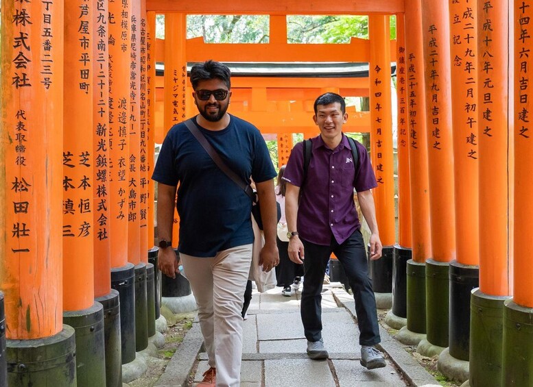 Two people walking through a corridor with orange pillars and Japanese writing on them. 1