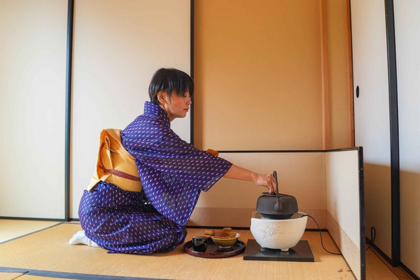 A person in traditional attire kneeling on tatami flooring, preparing matcha with a bowl and a whisk. 4