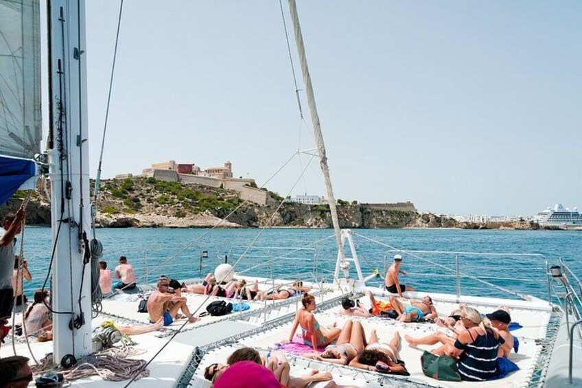 A sailboat with people sunbathing and swimming in the water, a castle in the background, and a clear blue sky. 2
