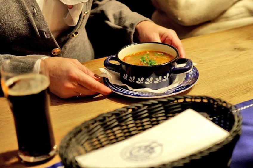 Une personne tenant un bol de soupe, accompagnée d’une tasse et d’une soucoupe à motifs, d’un verre contenant un liquide sombre et d’un panier avec une serviette. 3