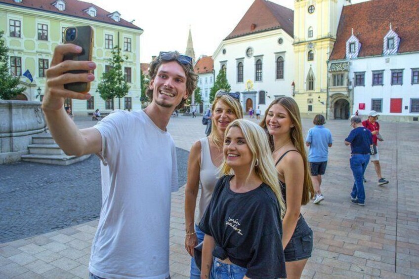 En grupp människor som tar en selfie på ett offentligt torg med historiska byggnader i bakgrunden. 4