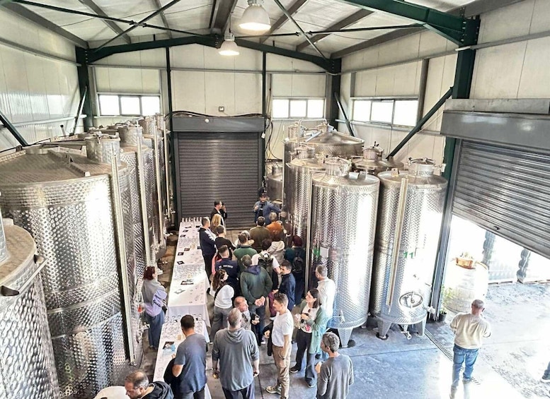 A group of people inside a large industrial building with stainless steel tanks and a table with items on display. 1