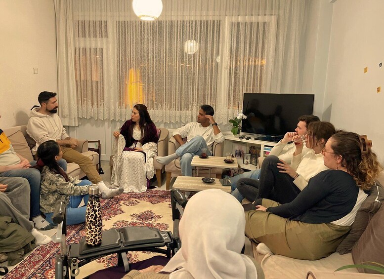 A group of people sitting on sofas and chairs in a living room with a television, a table with drinks, and a patterned rug. 1