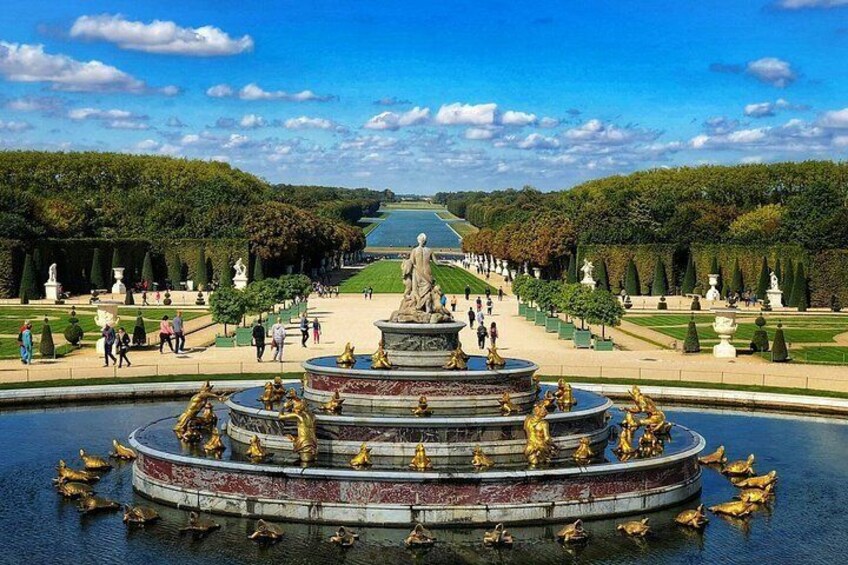 A grand fountain with statues, a long pathway lined with trees, and a body of water reflecting the sky. 4