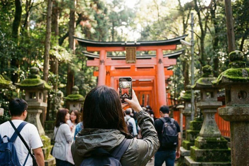 A person taking a photo with a smartphone, a red torii gate, and stone lanterns in a forested area. 2