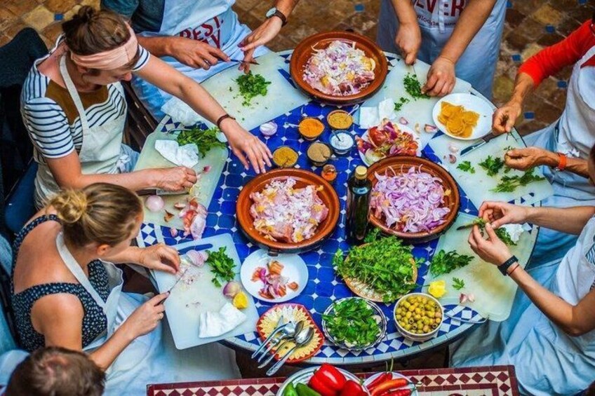 En grupp människor som lagar mat runt ett runt bord, med olika ingredienser och köksredskap utspridda. 2
