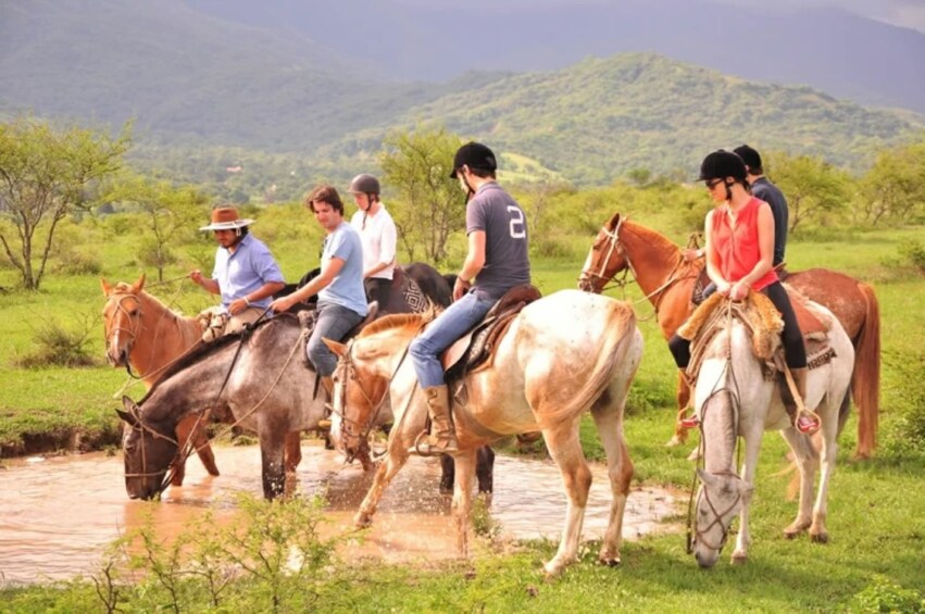 A group of people riding horses across a shallow water crossing in a grassy field with hills in the background. 1