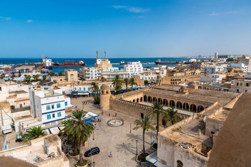 A coastal cityscape with a historic fort, palm trees, and a busy harbor. 1