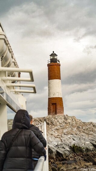 A person in a black jacket looking at a tall, striped lighthouse on a rocky shore. 3