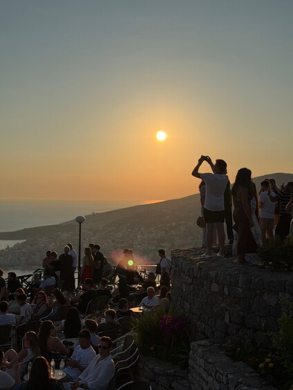 A sunset over a coastal city with people dining and standing on a stone platform. 2