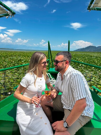 Two people sitting on a green platform in a field of water plants, holding drinks. 1