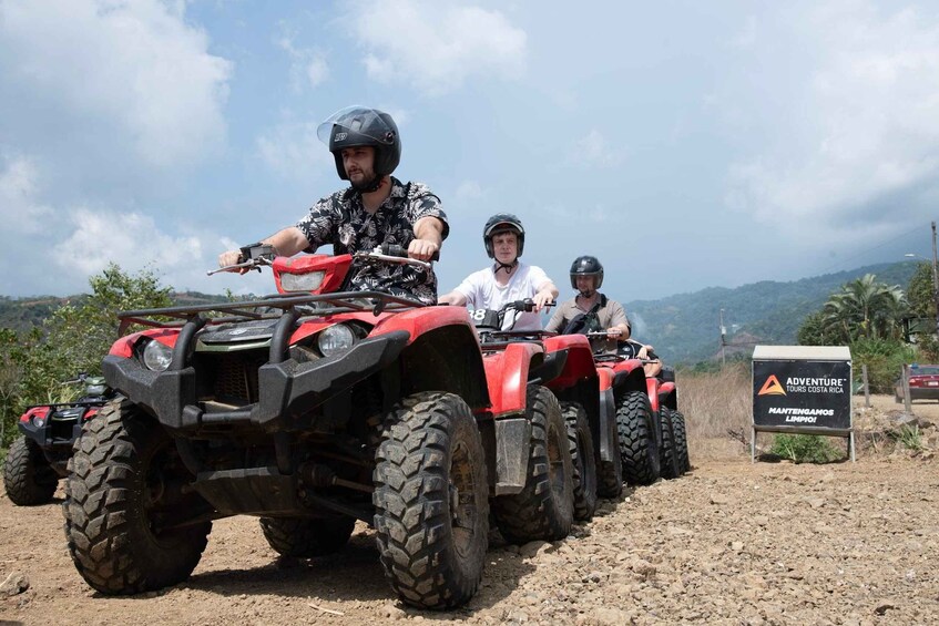 Een groep mensen op rode quadfietsen op een zandweg, met een bord voor Adventure Tours Costa Rica. 4