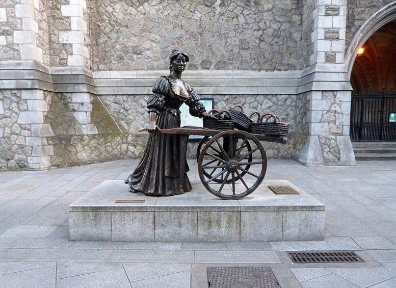 A statue of a woman in period clothing standing next to a wooden cart with baskets. 1