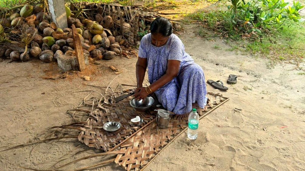 A person sitting on the ground, engaged in a food preparation activity, with a woven mat, metal bowls, and a plastic bottle nearby. 2