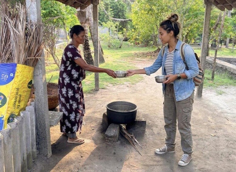 Two people exchanging bowls over a fire pit in a rural outdoor setting. 1