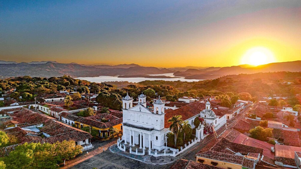 A white colonial-style church surrounded by terracotta-roofed buildings, set against a backdrop of mountains and a sunset over a lake. 1