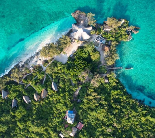 An aerial view of a tropical island with clear turquoise waters, dense green vegetation, and several small buildings. 5