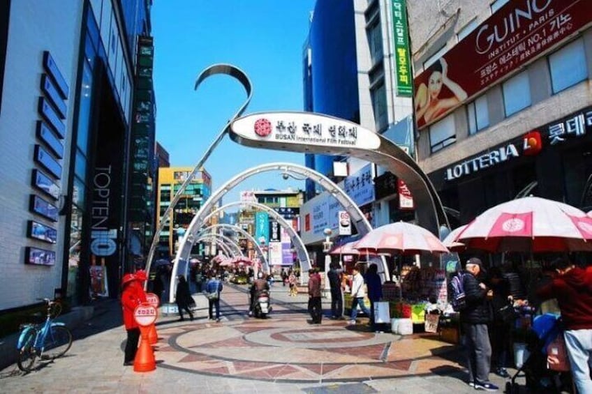 Una concurrida calle urbana con un gran arco, comercios y gente caminando. 1