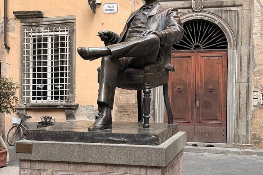 A bronze statue of a seated man with a cane, a bicycle, and a building with a wooden door and window in the background. 3