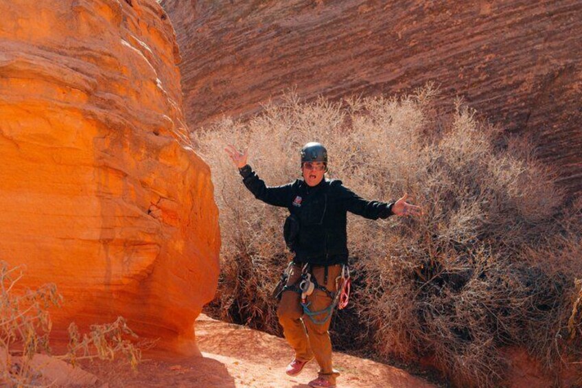 A person rappelling down a canyon with a red rock face and sparse vegetation. 2