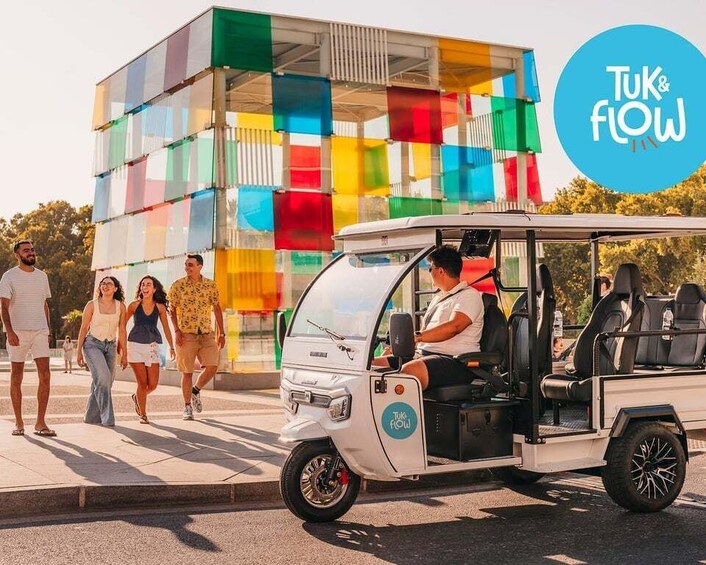 A tuk-tuk parked on a street with pedestrians walking by and a colorful building in the background. 1