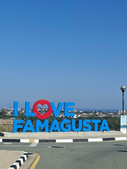 A sign with 'I LOVE FAMAGUSTA' in large blue letters, with a red heart in the middle, set against a clear blue sky and a distant cityscape. 2