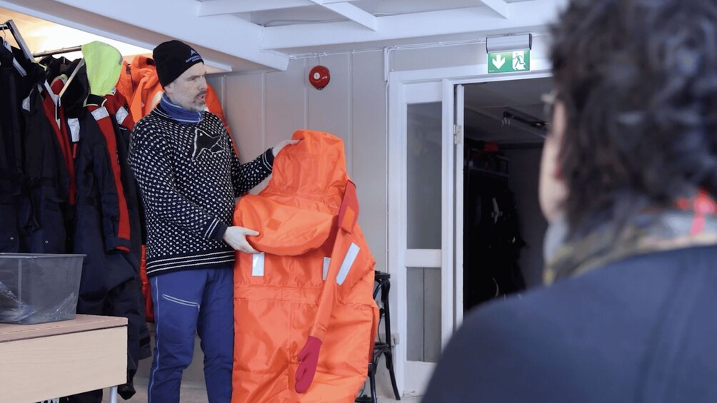 A person holding an orange jacket in a room with a white door and a red emergency exit sign. 2