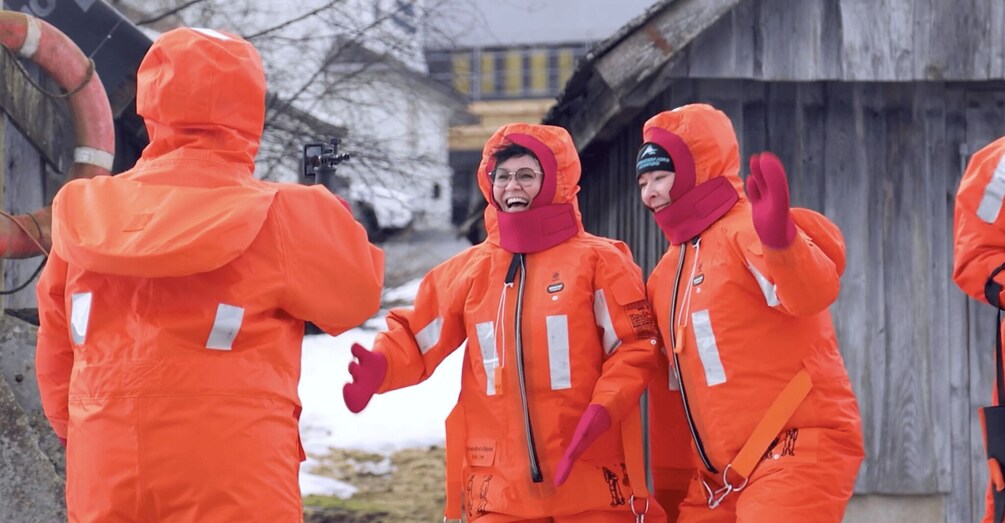 People in orange hazmat suits with reflective stripes, standing outdoors near a wooden structure. 4