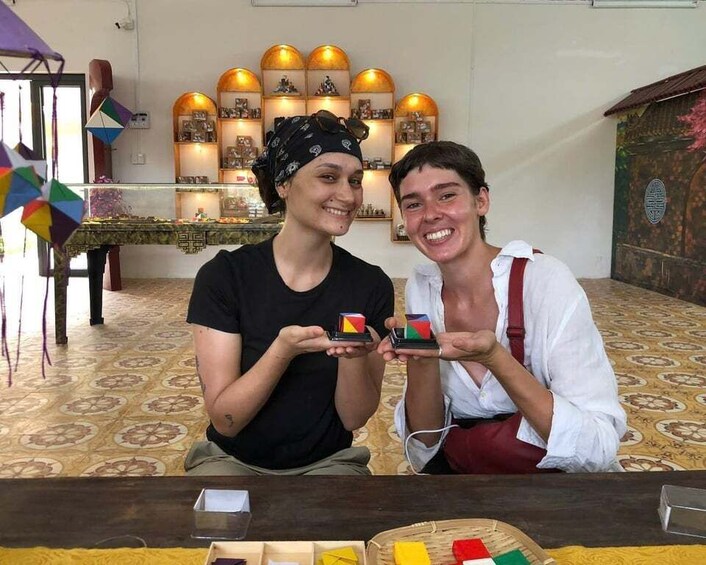 Dos personas sosteniendo objetos coloridos en una mesa en una habitación con piso estampado y estantes decorativos. 1