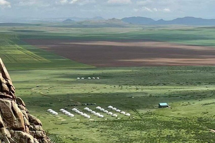 A vast landscape with green fields, a cluster of white tents, and distant mountains. 5