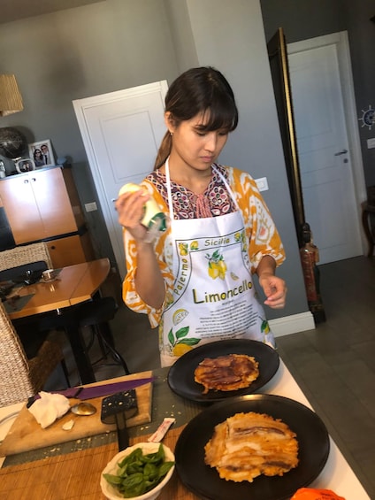 A person in a kitchen preparing food, wearing a 'Sicilia Limoncello' apron, with a cutting board, knife, and plates of food visible. 1