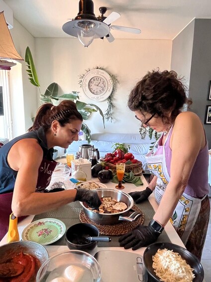 Two people preparing food in a kitchen with a clock on the wall, a ceiling fan, and various cooking utensils and ingredients on the counter. 1