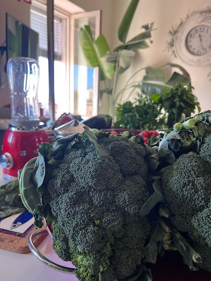 A kitchen counter with a red blender, a bowl of broccoli, and a window with a view of a building and plants. 3