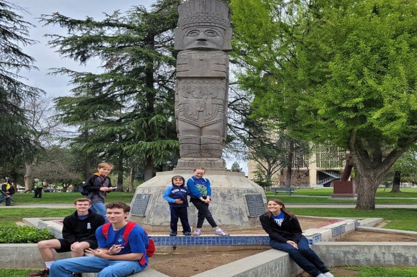 Un grupo de personas sentadas y de pie alrededor de un monumento de piedra en un parque con árboles y un edificio al fondo. 5